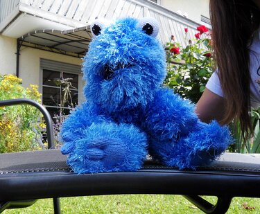 Handgestricktes, zotteliges blaues Plüschtier mit großen Augen auf einer Gartenliege.