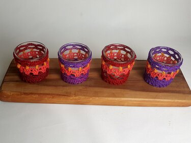 Vier Glas-Teelichthalter mit roten und lila gehäkelten Hüllen auf einem Holzbrett