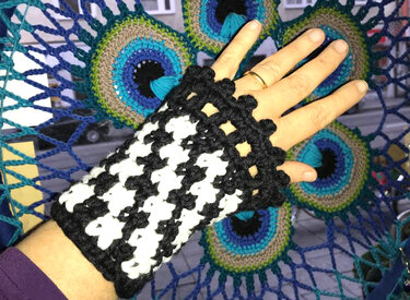 Hand mit schwarz-weißer, gehäkelter fingerloser Handschuh im Hahnentrittmuster vor gehäkeltem Hintergrund