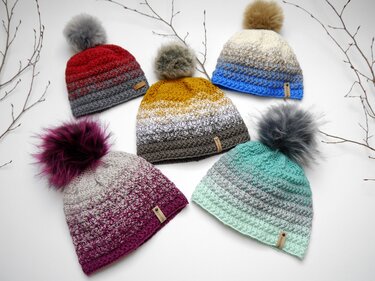 Fünf gehäkelte Bommelmützen in Rot, Gelb, Blau, Magenta und Mintgrün mit Farbverlauf und Bommeln auf hellem Hintergrund.