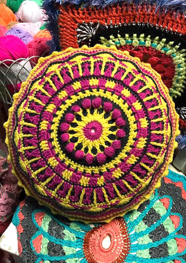 Detailaufnahme eines runden gehäkelten Kissens mit Noppenstich, gelben und pinken Ringen, weiterer Häkelware und Garn im Hintergrund