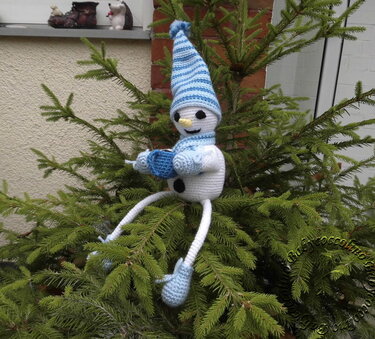 Häkel-Schneemann mit langen Beinen, blau-weiß gestreifter Mütze und Schal, sitzend in Tannenzweigen