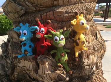 Vier gehäkelte Amigurumi-Giraffen in Blau, Rot, Grün und Gelb auf einem braunen Baumstamm.