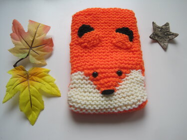 Gestrickte Fuchs‑Hülle in Orange mit weißer Schnauze, arrangiert neben bunten Herbstblättern und einem kleinen Stern aus Rinde
