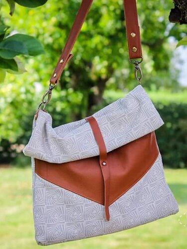 Helle Umhängetasche mit geometrischem Muster, V-förmigem braunen Besatz und Klappenverschluss, hängend an einem Baum