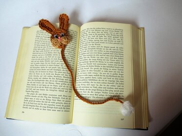 Gehäkeltes Hasen-Lesezeichen in Braun mit geflochtener Schnur und weißem Pompon auf offenem Buch