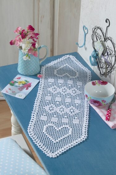 Romantischer weißer gehäkelter Läufer mit Herzmotiven auf blauem Tisch neben Blumen und Schale
