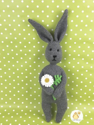 Grau gehäkelter Amigurumi-Hase mit langen Ohren, der eine weiße Margerite hält, vor grünem, gepunktetem Stoff