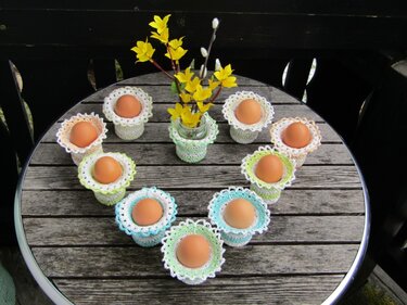 Neun gehäkelte Eierwärmer mit braunen Eiern in Herzform auf einem Holztisch, Vase mit gelben Frühlingsblüten