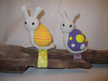 Zwei gehäkelte Amigurumi-Schnecken mit cremefarbenen Körpern, eine mit gelb-orangem gestreiftem Panzer, die andere mit violettem Panzer und Punkten, auf Treibholz