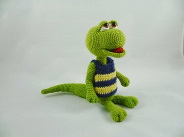 Gehäkelter grüner Amigurumi-Dinosaurier mit blau-gelb gestreiftem Shirt, großen Augen und roter Zunge, sitzt auf hellem Hintergrund.