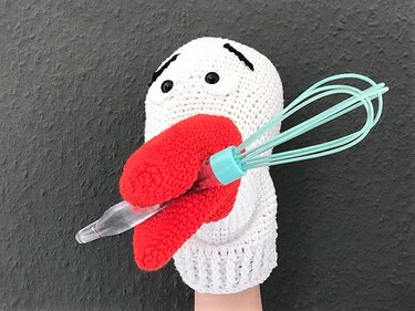 weiße gehäkelte Handpuppe mit roter Zunge hält türkisfarbenen Quirl vor dunklem Hintergrund