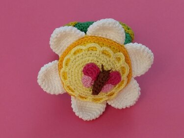 Handgehäkelte Blumenapplikation mit gelbem Zentrum und kleinem Schmetterling auf rosa Hintergrund