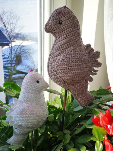 Zwei gehäkelte Amigurumi-Vögel, weißer und brauner, auf Stäben in einer Zimmerpflanze vor Fenster