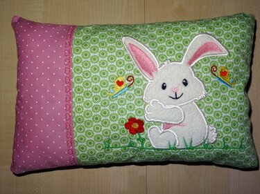 Stickerei eines freundlichen weißen Hasen, einer roten Blume und zweier Schmetterlinge auf hellgrün gemustertem Stoff neben rosa gepunktetem Stoff.