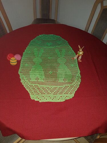 Ovales grünes Häkeldeckchen mit Hasenmotiv auf rotem Tisch, links drei bunte Eier, rechts kleine Hasenfigur.