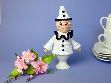 Gehäkelter Pierrot-Eierwärmer in Weiß und Schwarz auf einem Eierbecher, umgeben von rosa Blüten und weißem Geschirr.