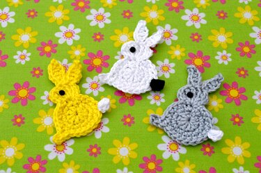 Drei gehäkelte Hasen-Applikationen in Gelb, Weiß und Grau auf grünem Blumenstoff
