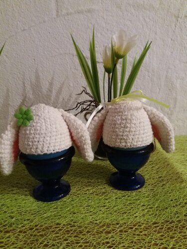 Zwei weiße gehäkelte Hasen-Eierwärmer mit Schlappohren in dunkelblauen Eierbechern auf grünem Tuch, im Hintergrund künstliche Blumen.