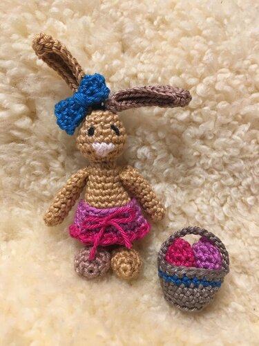 Gehäkelter brauner Amigurumi-Hase mit blauer Schleife, rosa Rock und kleinem Korb mit bunten Eiern auf hellem flauschigem Stoff