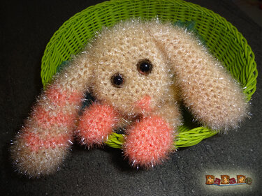 Gehäkeltes flauschiges Häschen in hellbeige mit rosa Details sitzt in einem grünen Korb.