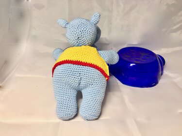Rückansicht: gehäkeltes hellblaues Flusspferd-Amigurumi mit gelbem Shirt und rotem Saum neben blauer Schale