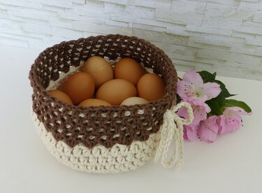 Gehäkelter Korb in Braun und Creme, gefüllt mit braunen Eiern, neben rosa Blüten