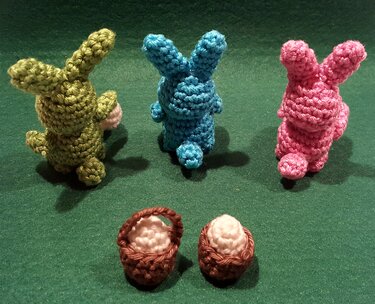 Drei gehäkelte Amigurumi-Hasen (grün, blau, rosa) mit zwei kleinen braunen Körbchen auf grünem Untergrund.