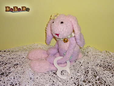 Gehäkeltes rosa-lila Amigurumi-Häschen mit Glöckchen und weißem Beißring auf weißer Häkeldecke.