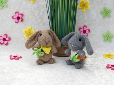 Zwei gehäkelte Hasen (braun und grau) mit Halstüchern, Karotte und Blatt, umgeben von Filzblumen auf hellem Untergrund.