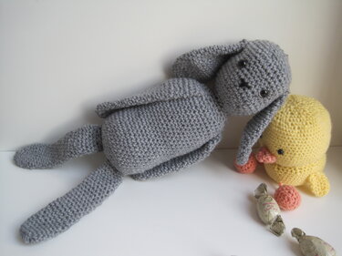Grau gehäkelter Amigurumi-Hase neben gelbem Häkelküken auf weißem Untergrund; Bonbons im Vordergrund.