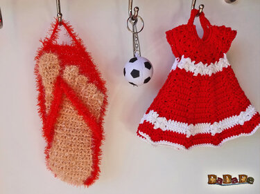 Zwei gehäkelte Anhänger an Haken: rotes Mini-Kleid mit weißen Streifen und beige-rote flauschige Badelatsche; dazwischen kleiner Fußball‑Schlüsselanhänger.