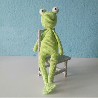 Hellgrüner gehäkelter Frosch-Amigurumi mit großen Augen auf kleinem gepunkteten Stuhl.