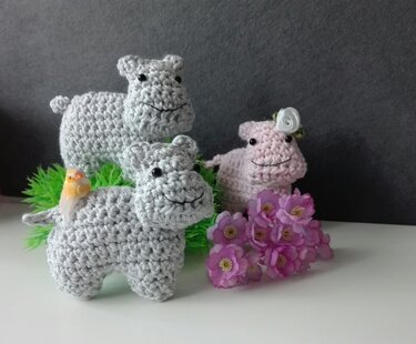 Drei gehäkelte Nilpferd-Amigurumi, zwei grau und eins rosa, mit schwarzen Augen und dekorativen Blumen