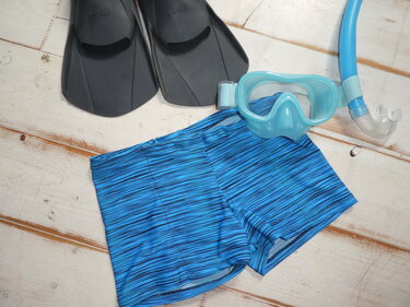 Blaue gestreifte Kinder-Badehose neben schwarzen Schwimmflossen und hellblauer Tauchmaske mit Schnorchel auf Holz