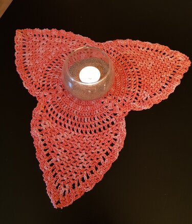 Korallenrotes, dreiblättriges gehäkeltes Deckchen mit Glas-Teelichthalter auf dunklem Untergrund.