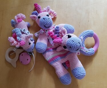 Gehäkeltes Baby-Set mit Einhorn-Motiv: Ein Schmusetuch, eine Schnullerkette mit Buchstaben und ein Greifling in Rosa, Lila und Hellblau auf einem Holztisch