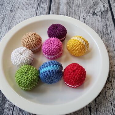 Acht bunte, gehäkelte Macarons auf einem weißen Teller vor Holzoptik-Hintergrund