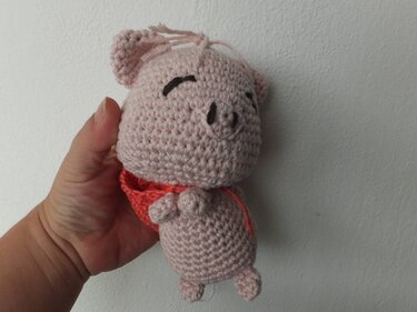 Hand hält ein gehäkeltes rosa Amigurumi‑Schwein mit rotem Halstuch vor heller Wand