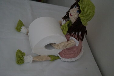Handgehäkelte Amigurumi-Puppe in rosa Kleid und grüner Mütze, die eine Toilettenpapierrolle hält.
