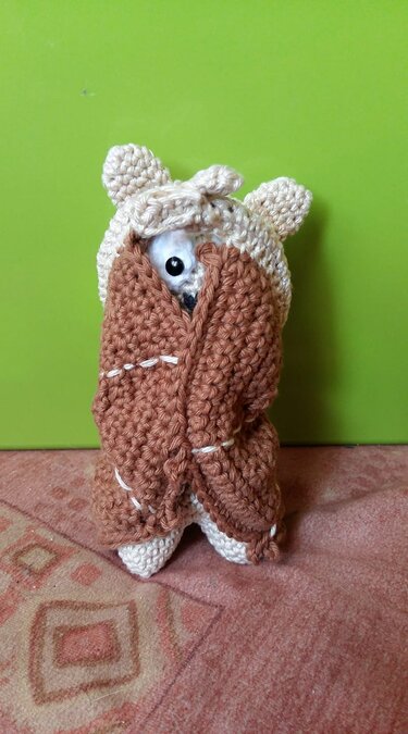 Gehäkelte Amigurumi-Figur in beigefarbener Kapuze mit Ohren, im braunen Wickelmantel ein Auge blickt hervor