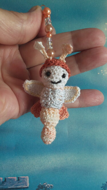 Hand hält kleines gehäkeltes Insekten-Amigurumi in Pfirsich- und Cremetönen mit Perlenaufhängung