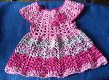 Gehäkeltes Babykleid in drei Rosa- und Pinktönen mit verschiedenen Musterbereichen und applizierten Häkelblumen, liegend auf blauem Stoff.