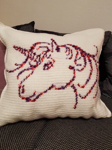 Weißes gehäkeltes Dekokissen mit Einhorn‑Umriss in Rot-, Pink- und Violetttönen auf einer dunklen Couch