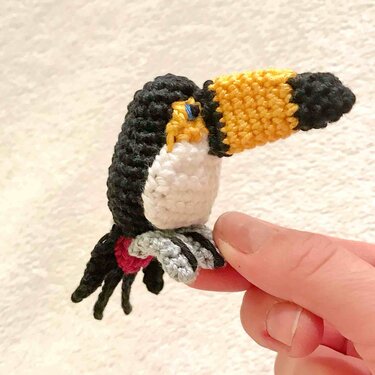 Mini‑gehäkelter Tukan (Amigurumi) mit gelb‑schwarzem Schnabel, zwischen zwei Fingern gehalten