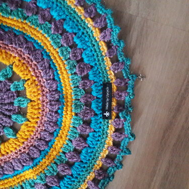 Rundes Häkel-Mandala in Türkis, Gelb und Lila mit Noppen- und Muschelmuster auf Holzuntergrund