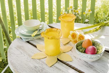 Gelbe Stoff-Windlichter auf blütenförmigen Untersetzern auf Holztisch mit Narzissen und bunten Ostereiern