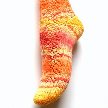 Gestrickte Damen‑Socke in Gelb, Orange und Rot mit feinem Lochmuster vor hellem Hintergrund.