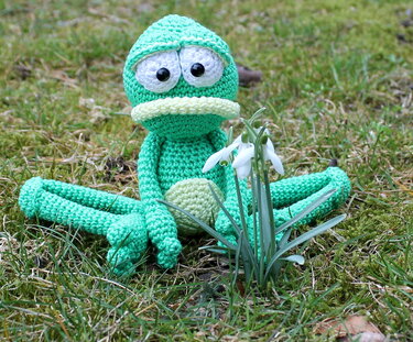 Gehäkelter grüner Amigurumi-Frosch neben Schneeglöckchen im Gras