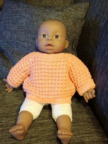 Nahaufnahme einer Babypuppe mit handgehäkeltem lachsfarbenem Pullover und weißer Hose